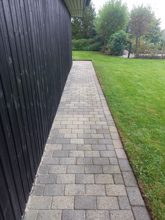 En lige flisegang langs et sortmalet hus, flankeret af veltrimmet græsplæne og buske. Et eksempel på elegant og praktisk brolægning.